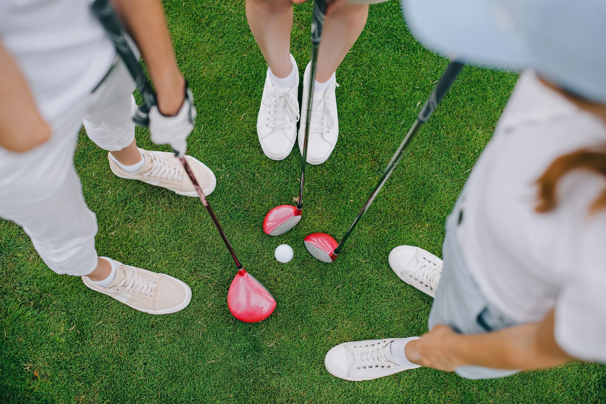 overhead view of female golf players with golf clubs standing on green lawn with golf ball in middle